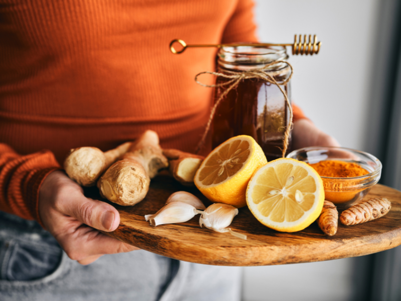 Life Extension europe: Person wearing orange sweater and gre pants, carrying a wooden tray with antioxidant ingredients. Lemon, garlic, tumeric.