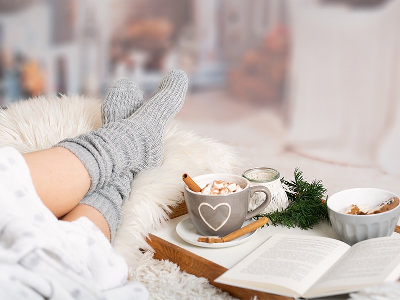 Life Extension, relaxed woman cuddled up on the couch enjoying a cozy heart mug with a warm drink, cookies, and a book