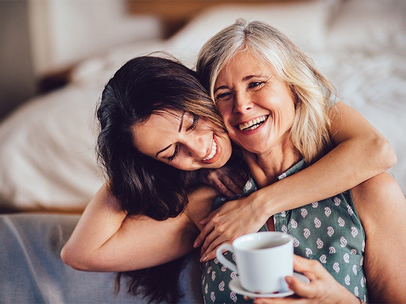 Life Extension, vital looking gray haired, smiling woman enjoying her warm drink and being hugged by her dark haired daughter
