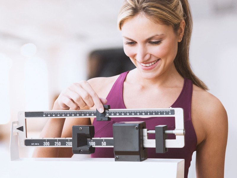 Life Extension, blonde woman in purple top smiling while measuring her body weight